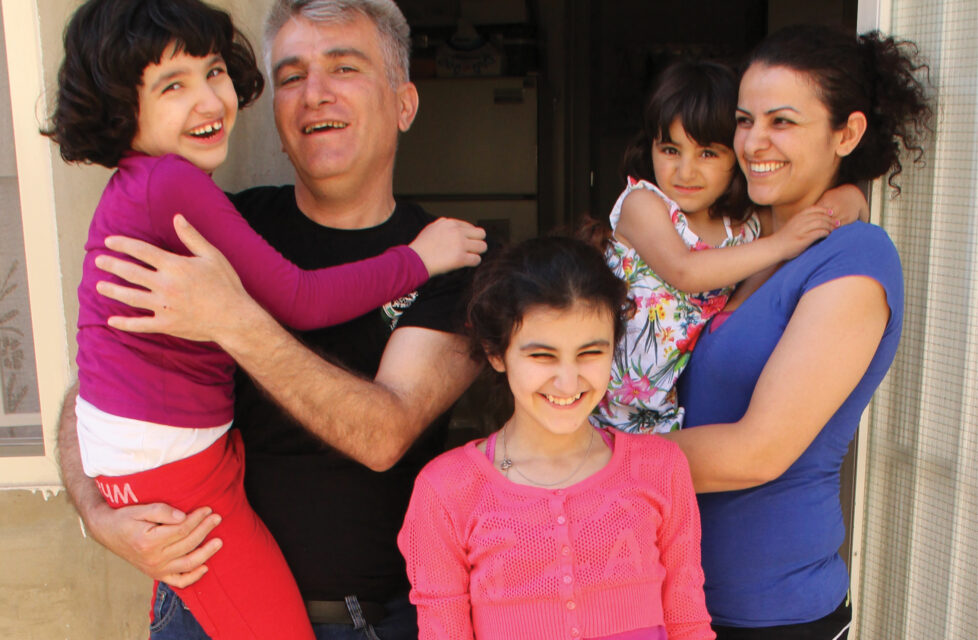a husband and wife stand with their three girls outside their house smiling